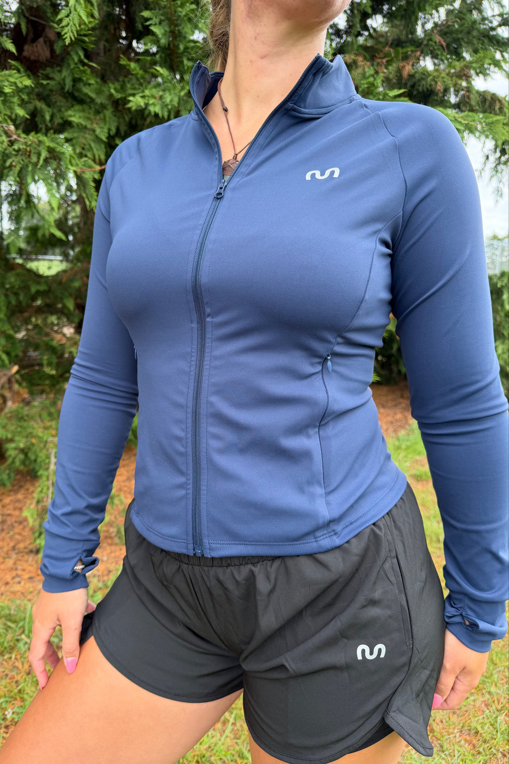 Women wearing a blue jacket and gray shorts outdoors with greenery in the background