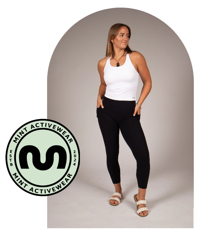 Woman wearing a white tank top and black leggings standing in front of a gray archway with a brand logo.